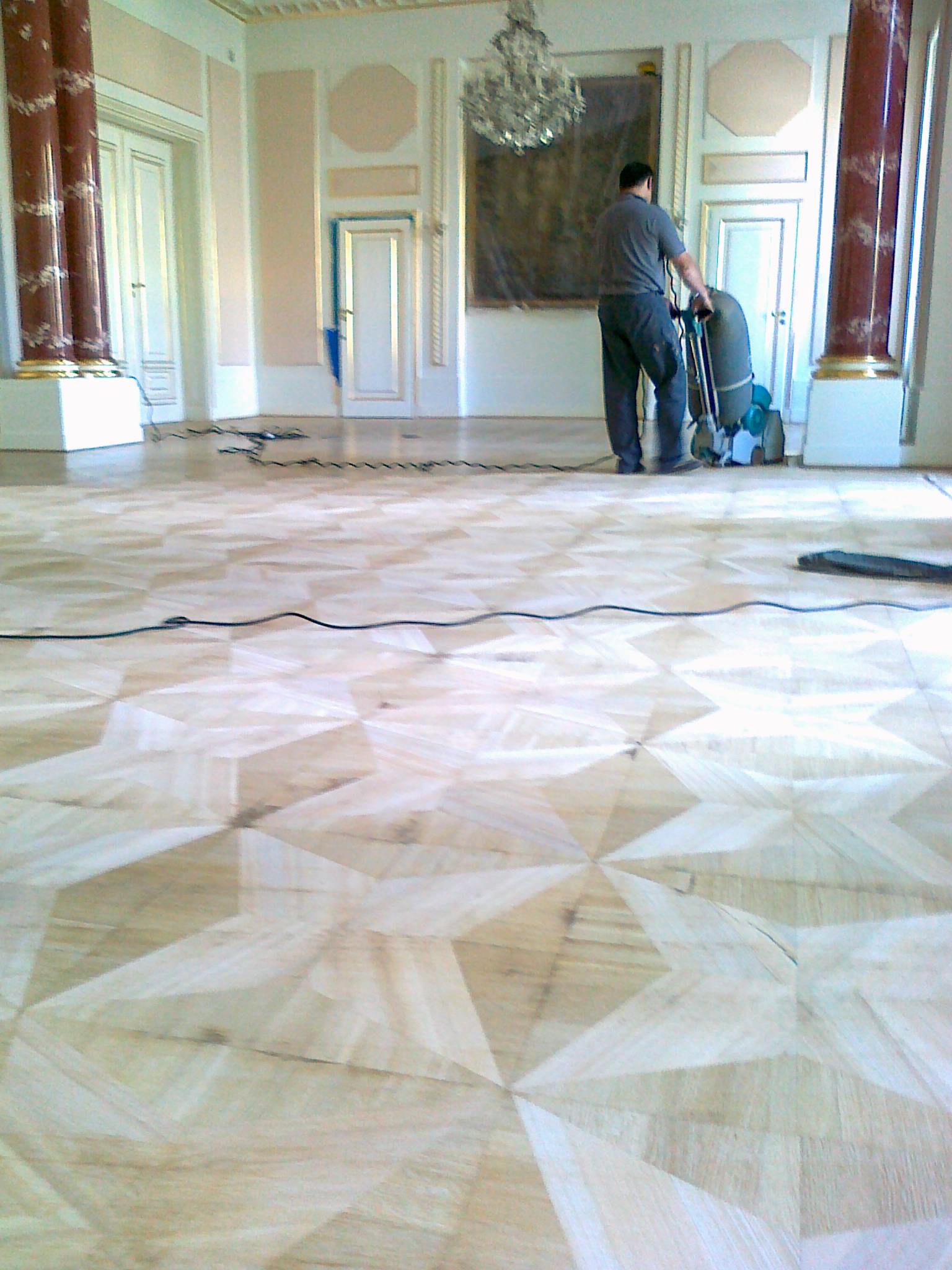 Parkiet w jodełkę podczas cyklinowania w przestronnym wnętrzu z kolumnami i żyrandolem, widoczna maszyna do cyklinowania i kabel zasilający.