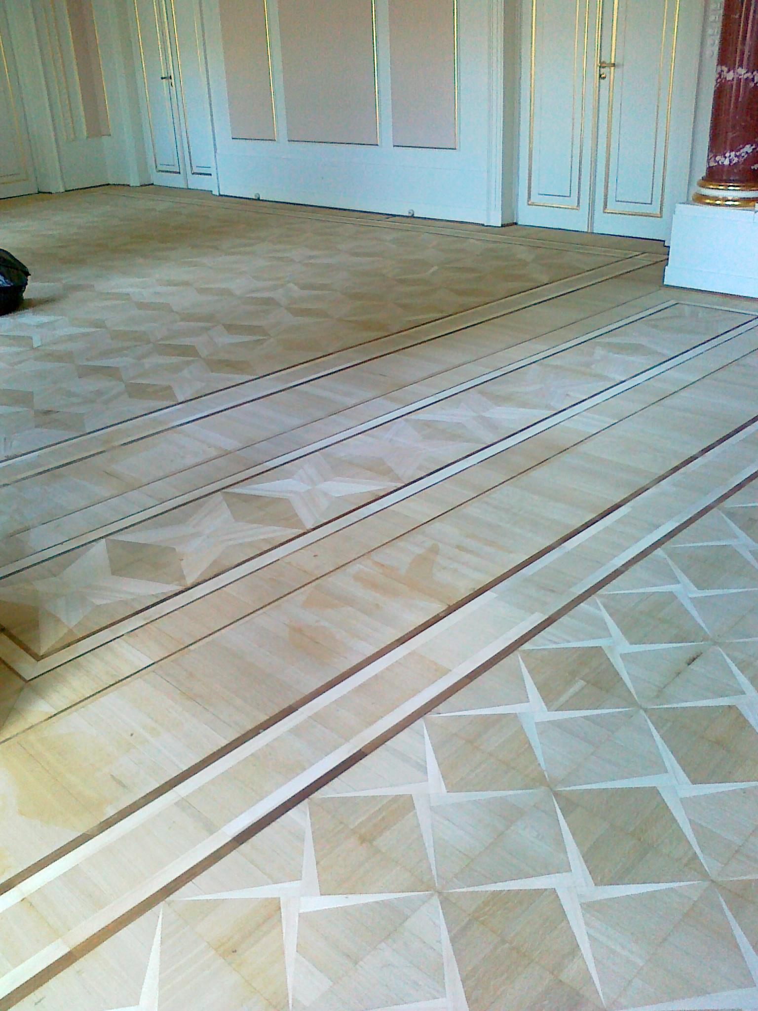 Wzór parkietu po renowacji w jasnych odcieniach drewna, z dekoracyjnymi bordiurami i geometrycznymi elementami, widoczne drzwi z ozdobnymi listwami i fragment kolumny w tle.