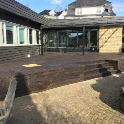 Ciemnobrązowy taras z drewna z niskimi schodami, przylegający do domu z szarymi ścianami i białymi oknami, widok z poziomu brukowanego patio.