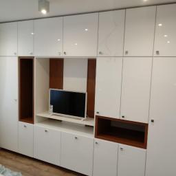 MM-Design - Biała, lakierowana zabudowa ścienna z wnęką na telewizor i półkami z ciemnego drewna, widoczna część okna z zasłoną.