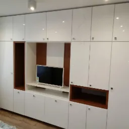 Biała, lakierowana zabudowa ścienna z wnęką na telewizor i półkami z ciemnego drewna, widoczna część okna z zasłoną.