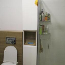 MM-Design - Biała szafka łazienkowa z otwartą półką, umieszczona obok toalety, z widocznymi kosmetykami na szklanych półkach w kabinie prysznicowej i żółtą rękawicą wiszącą na górze.