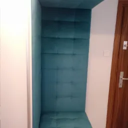 Wnęka z siedziskiem i tapicerowanym panelem ściennym w kolorze turkusowym, wkomponowana w białą szafę, obok drewnianych drzwi.