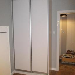 MM-Design - Biała szafa wnękowa z przesuwnymi drzwiami i srebrnymi obramowaniami, w rogu pokoju z drewnianą podłogą i widocznym wejściem do innego pomieszczenia.