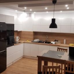 MM-Design - Nowoczesna kuchnia z białymi szafkami, drewnianym blatem i czarnym sprzętem AGD, oświetlona dwoma czarnymi lampami wiszącymi.