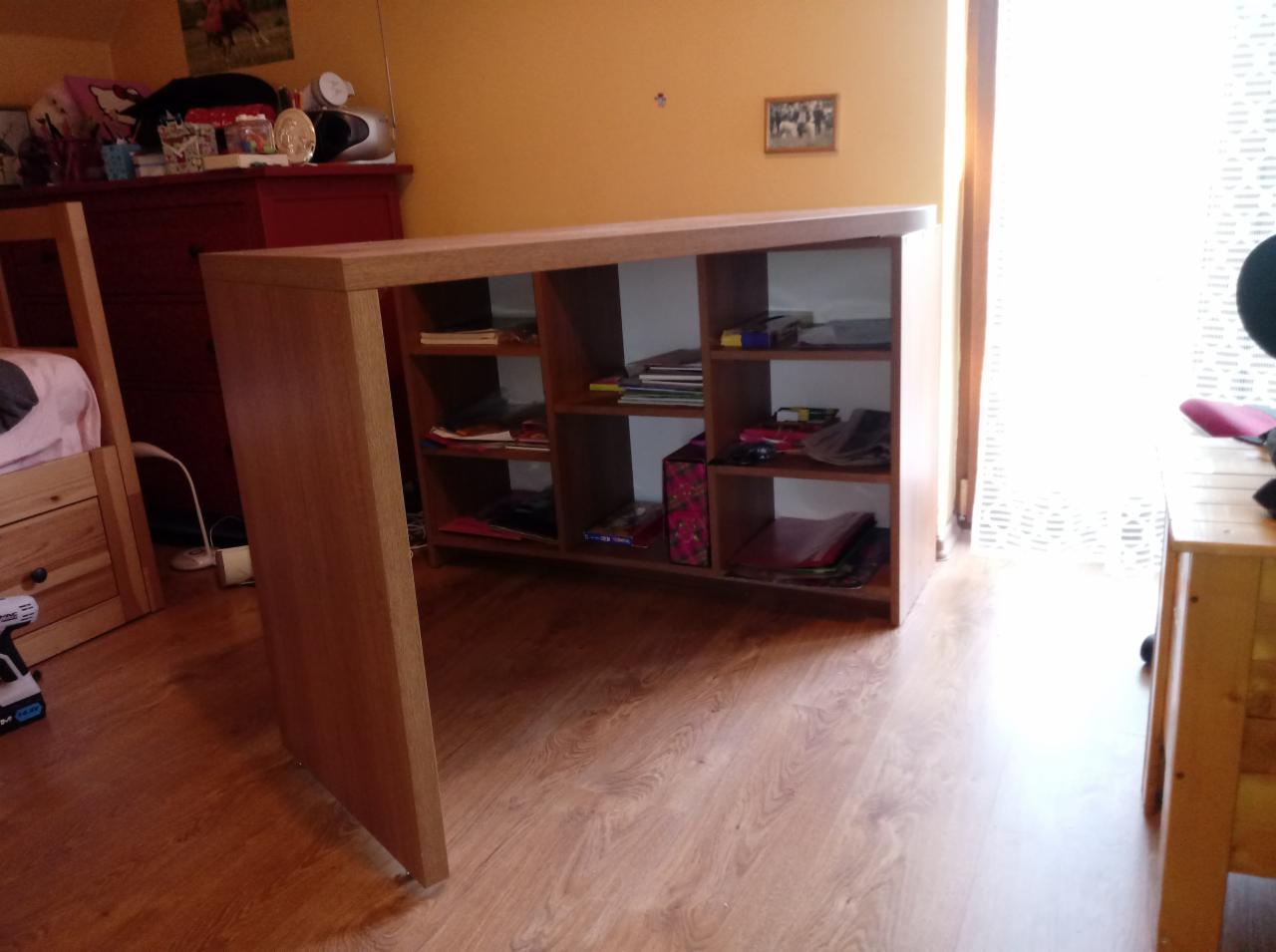 Drewniane biurko z otwartymi półkami, ustawione w pokoju z drewnianą podłogą, widok na wnętrze półek wypełnionych książkami i akcesoriami.