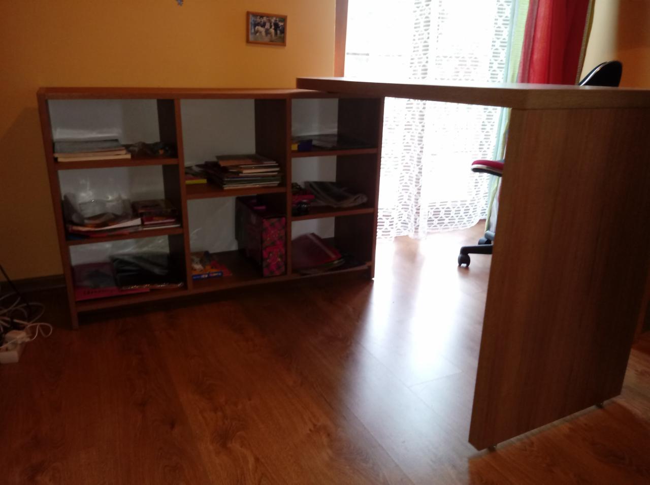 Biurko z obracanym blatem, połączone z regałem na książki, na podłodze panele drewnopodobne, w tle okno z zasłonami.