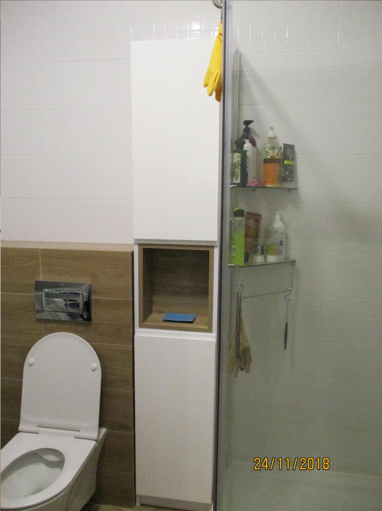 Biała szafka łazienkowa z otwartą półką, umieszczona obok toalety, z widocznymi kosmetykami na szklanych półkach w kabinie prysznicowej i żółtą rękawicą wiszącą na górze.