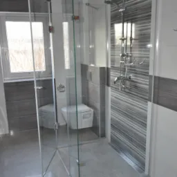 Nowoczesna kabina prysznicowa z transparentnego szkła, z deszczownicą i odpływem liniowym w posadzce, widok na toaletę i okno.