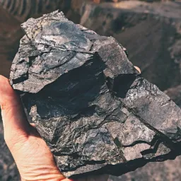 Dłoń trzyma bryłę węgla kamiennego na tle hałdy z widocznymi oponami ciężkiego sprzętu.