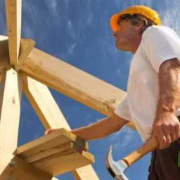 Monter w żółtym kasku mocuje drewnianą konstrukcję dachu na tle błękitnego nieba, widoczne narzędzia i logo firmy Bau-Tech.