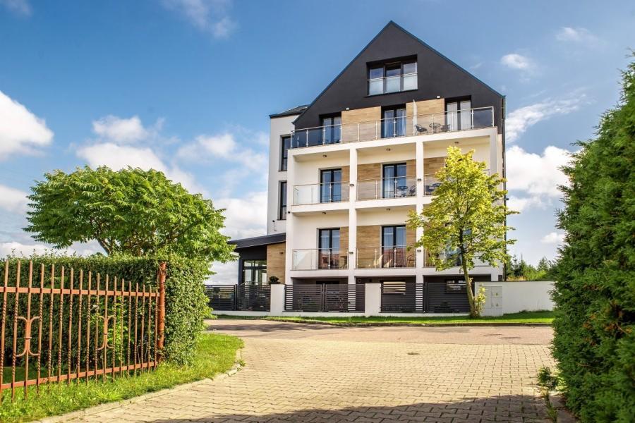 Nowoczesny, trzypiętrowy budynek hotelowy z balkonami i szklanymi balustradami, otoczony zielenią i ogrodzeniem.
