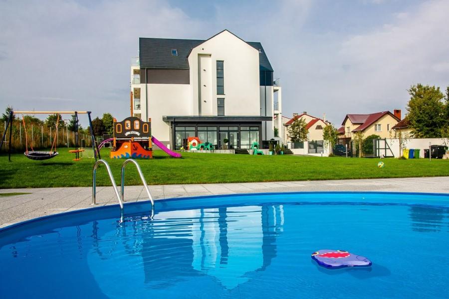 Nowoczesny hotel z basenem zewnętrznym, placem zabaw i huśtawkami na zielonym trawniku.