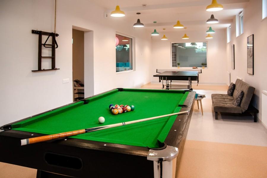 Pokój rekreacyjny z zielonym stołem bilardowym, kijem, kulami, stołem do ping-ponga i szarą sofą pod ścianą, oświetlony kolorowymi lampami.