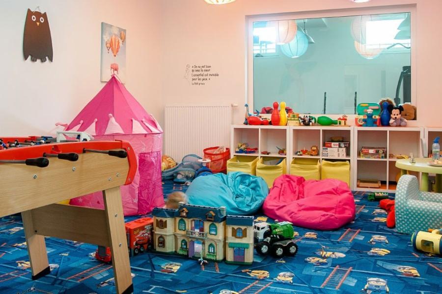 Pokój zabaw dla dzieci z piłkarzykami, różowym namiotem tipi, regałami z zabawkami, pufami w kolorach niebieskim i różowym oraz dywanem z motywem samochodów.