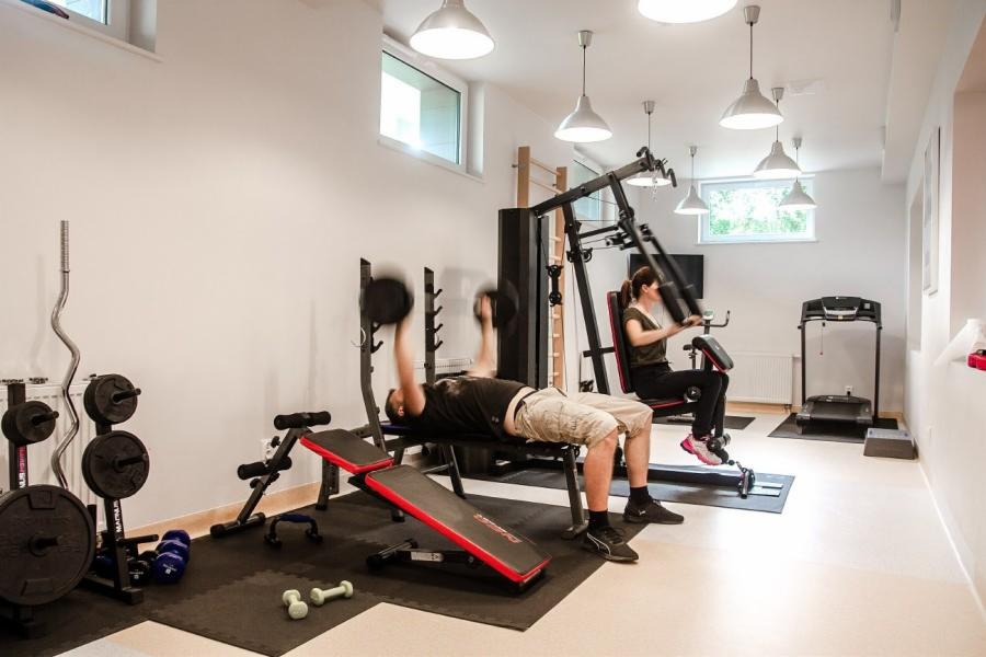 Domowa siłownia z mężczyzną wyciskającym sztangielki na ławeczce i kobietą ćwiczącą na maszynie, w tle bieżnia i drabinka gimnastyczna.