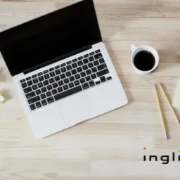 Widok z góry na otwarty laptop, kubek z kawą, notatki, ołówki i zmięte kartki na jasnym drewnianym biurku z widocznym logo 'inglitin' w prawym dolnym rogu.