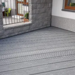 Szara deska tarasowa kompozytowa, widok z narożnika, z fragmentem muru z szarej cegły i elewacji budynku z oknem.