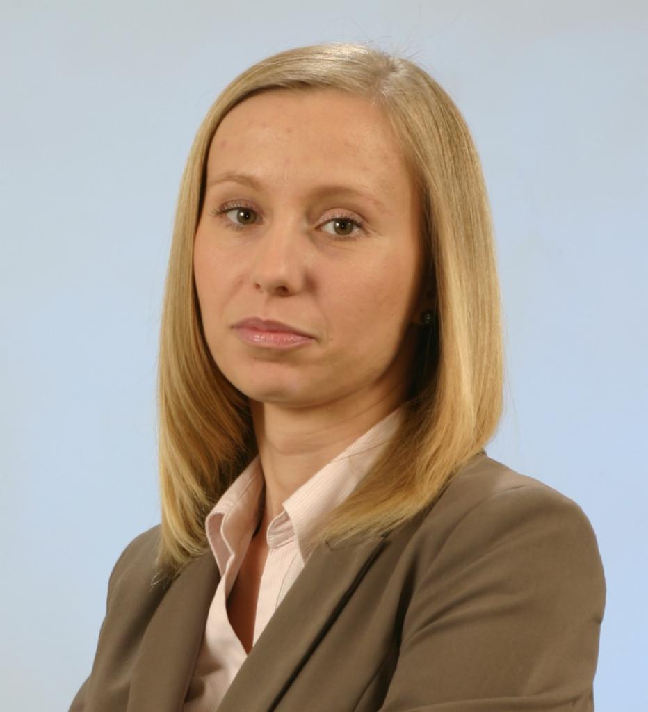 Portret kobiety z blond włosami w brązowej marynarce i bladoróżowej koszuli na jasnoniebieskim tle.