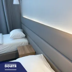 Pokój hotelowy z dwoma łóżkami, szafką nocną i podświetlanym zagłówkiem LED, widoczny fragment sufitu z lampą wiszącą i zasłona.