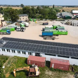 Widok z góry na budynek z panelami słonecznymi na dachu, teren firmy recyklingowej z kontenerami, ciężarówkami i stertami sprasowanego papieru.