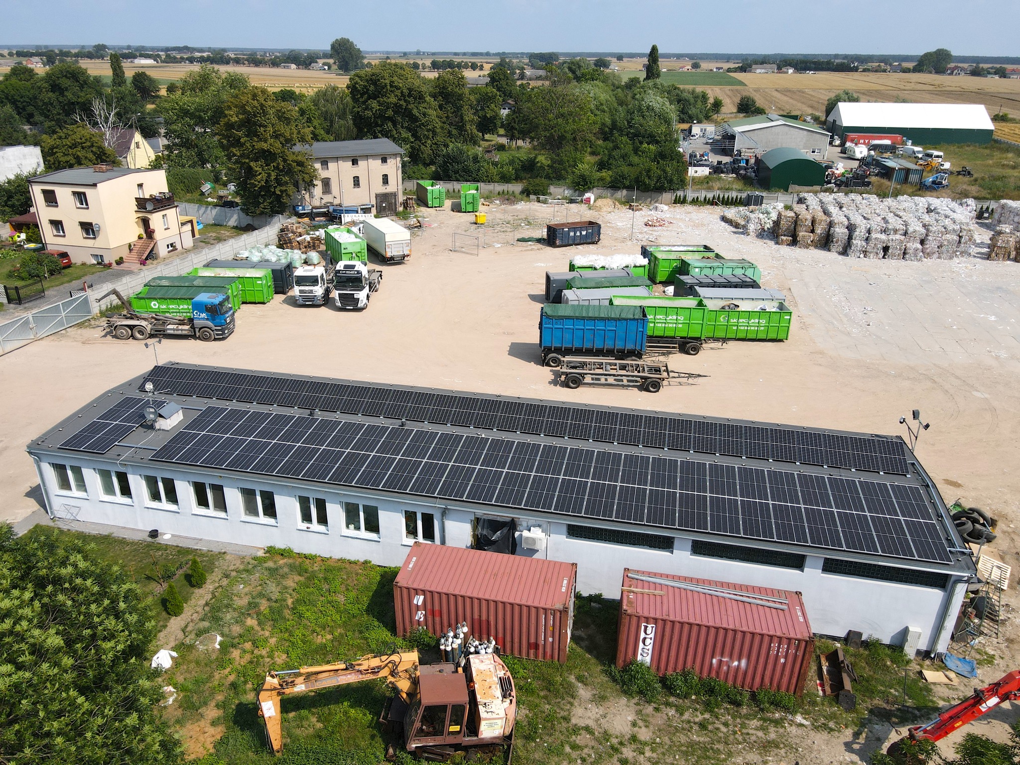 Widok z góry na budynek z panelami słonecznymi na dachu, teren firmy recyklingowej z kontenerami, ciężarówkami i stertami sprasowanego papieru.