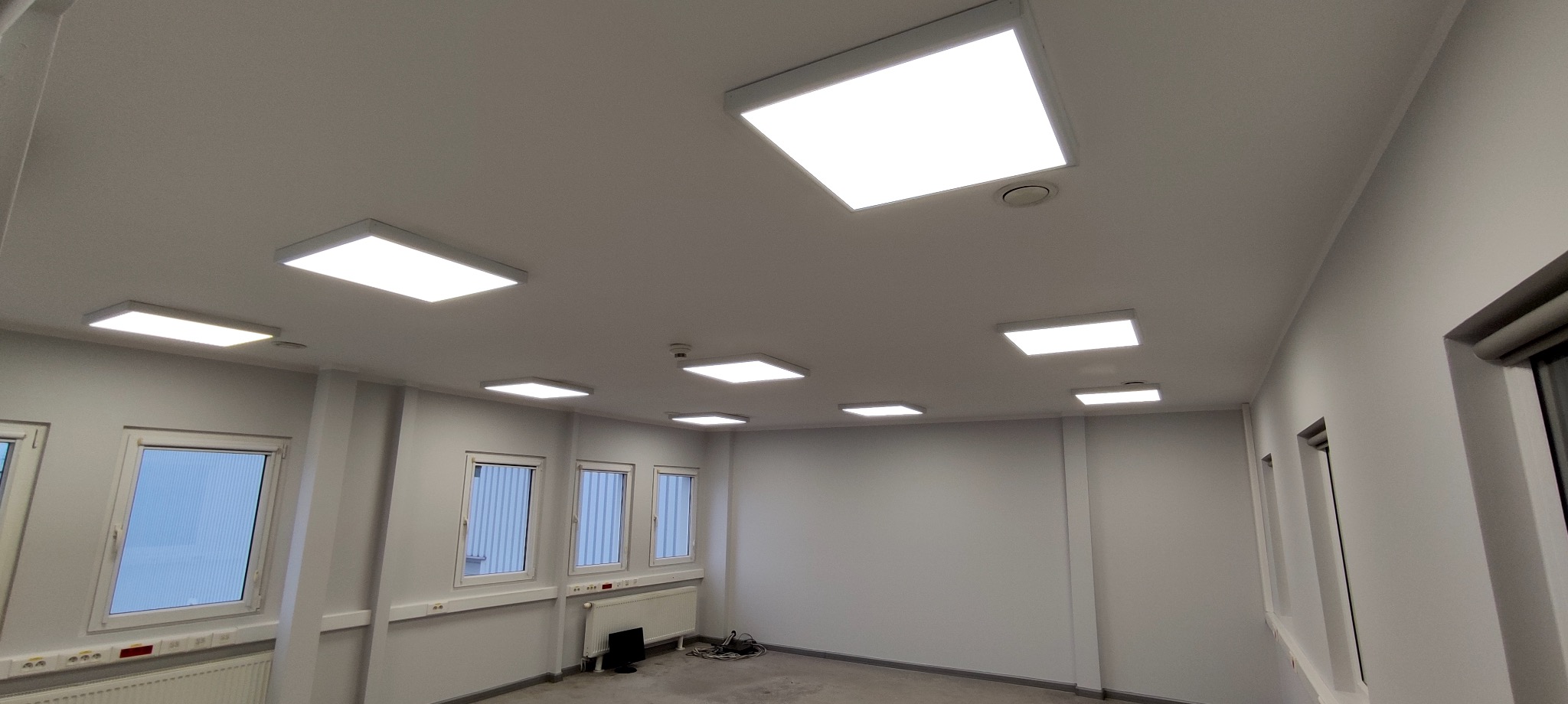 Białe, prostokątne panele LED zamontowane na suficie w jasnym pomieszczeniu biurowym z oknami.