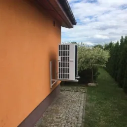 Biała jednostka zewnętrzna pompy ciepła zamontowana na pomarańczowej ścianie budynku, widok z boku na tle zieleni i pochmurnego nieba.