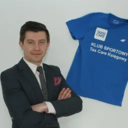 Mężczyzna w garniturze z poszetką w kratę na tle niebieskiej koszulki sportowej z napisem 'KLUB SPORTOWY Tax Care Księgowy' i logo firmy.