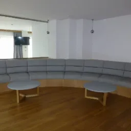 Długa, szara, tapicerowana sofa w kształcie łuku z drewnianą podstawą, uzupełniona dwoma okrągłymi stolikami kawowymi na drewnianych nogach, na tle parkietu i białych ścian z widocznym fragmentem...