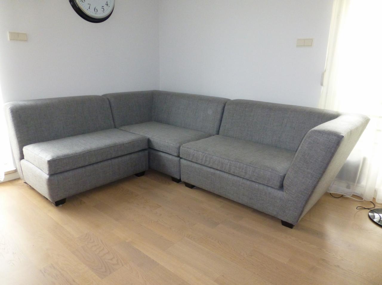 Szara, modułowa sofa narożna z tkaniny o wyraźnym splocie, ustawiona na drewnianej podłodze w jasnym pomieszczeniu z zegarem na ścianie.