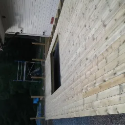 Drewniana elewacja domu w trakcie budowy, widoczne deski elewacyjne, otwór okienny, rusztowanie i drabina w tle.