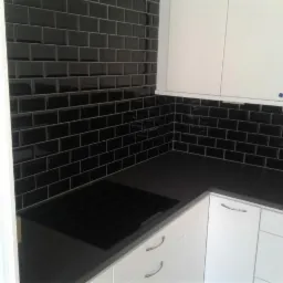 Błyszczące, czarne płytki ułożone w cegiełkę na ścianie nad blatem kuchennym, białe szafki z metalowymi uchwytami, czarna płyta indukcyjna.