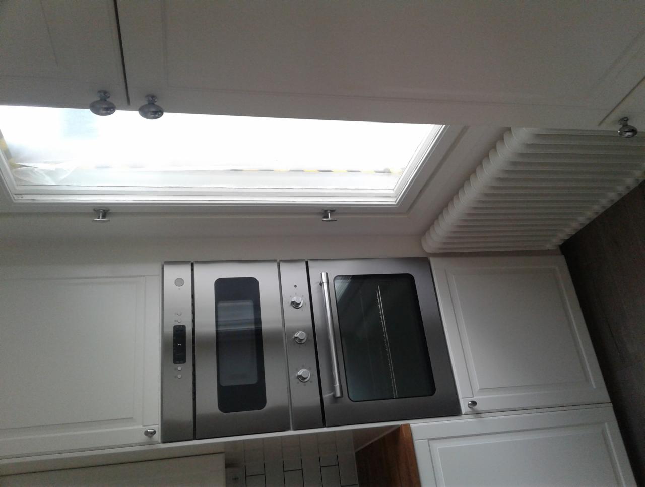 Nowoczesna zabudowa kuchenna z podwójnym piekarnikiem ze stali nierdzewnej, białymi szafkami i oknem dachowym z roletą plisowaną.