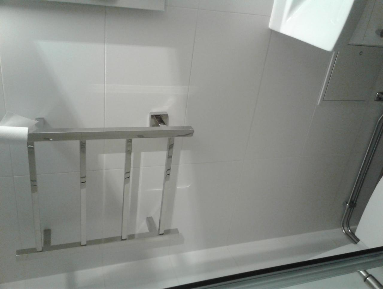 Nowoczesna, chromowana drabinka na ręczniki zamontowana na białych, kwadratowych płytkach w łazience, widoczny fragment umywalki i rury odpływowej.