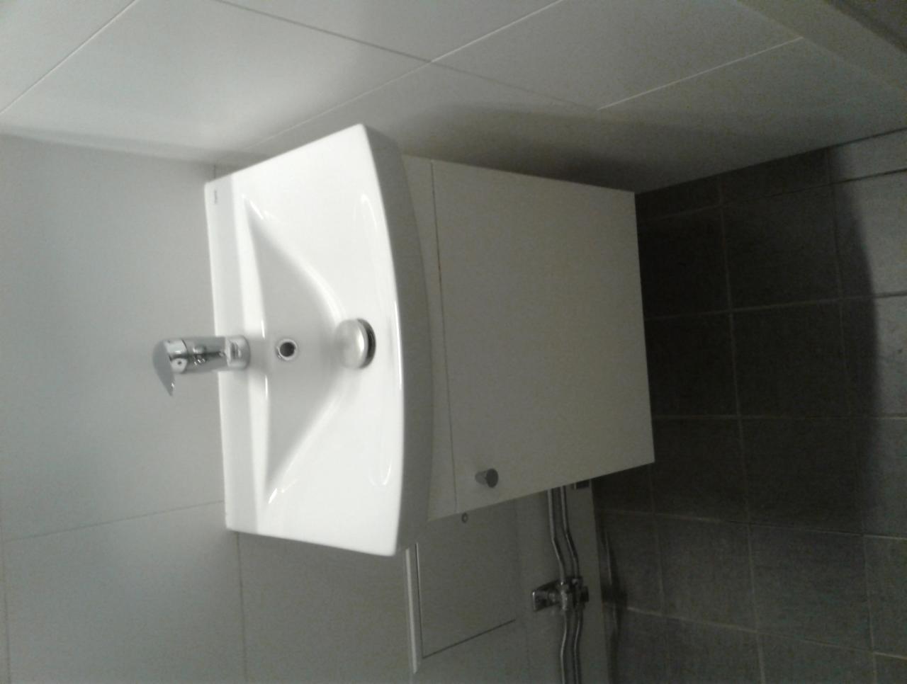 Biała umywalka z szafką wiszącą na ścianie z szarymi płytkami, widoczny chromowany kran i syfon