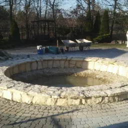 Okrągły, kamienny zbiornik z wodą w trakcie budowy, otoczony kostką brukową, z narzędziami i materiałami budowlanymi w tle.