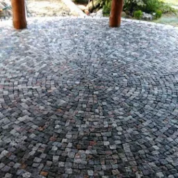 Okrągła nawierzchnia z drobnej, szarej kostki ułożonej koncentrycznie wokół dwóch drewnianych słupów, z zielenią w tle.