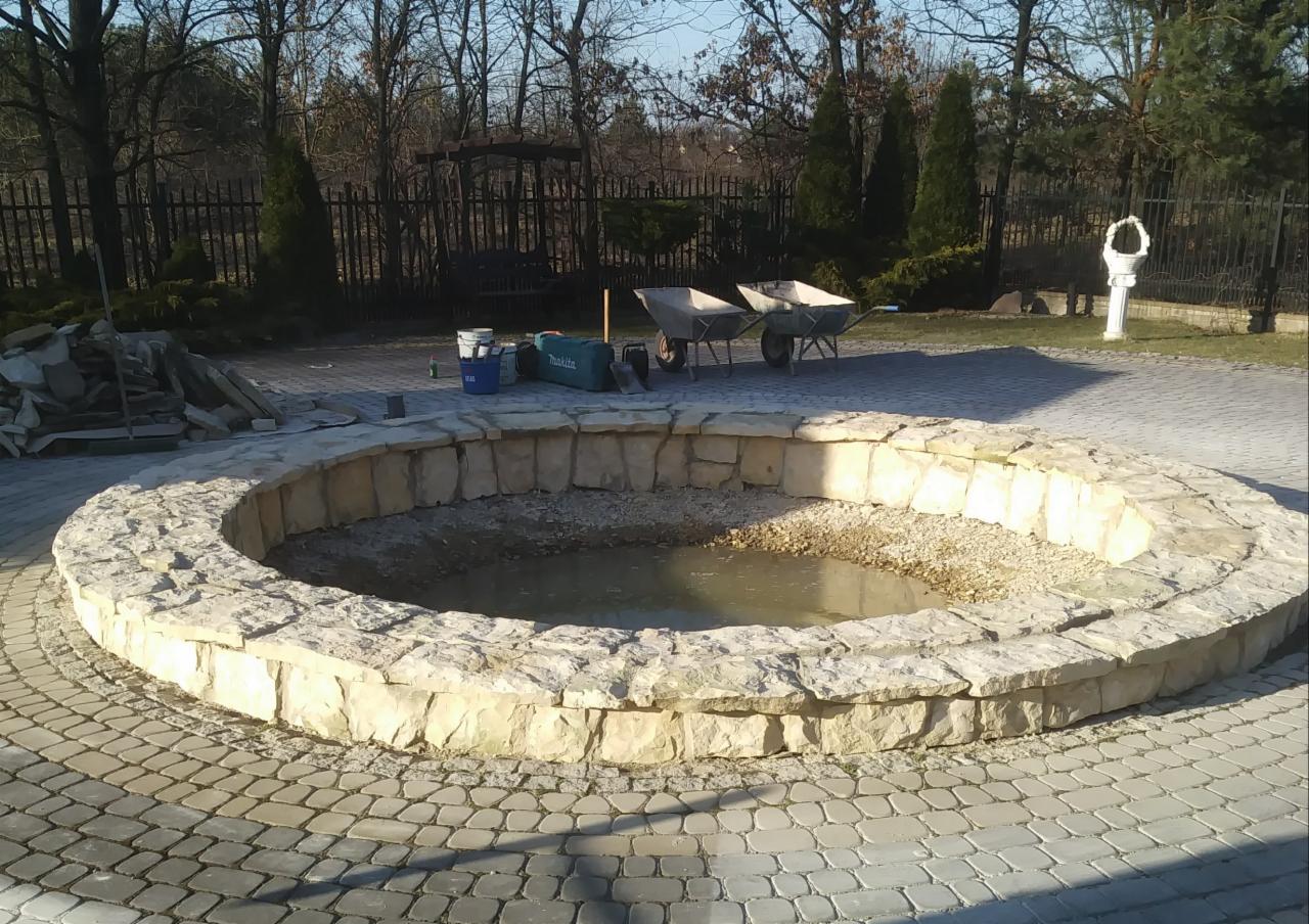 Okrągły, kamienny zbiornik z wodą w trakcie budowy, otoczony kostką brukową, z narzędziami i materiałami budowlanymi w tle.