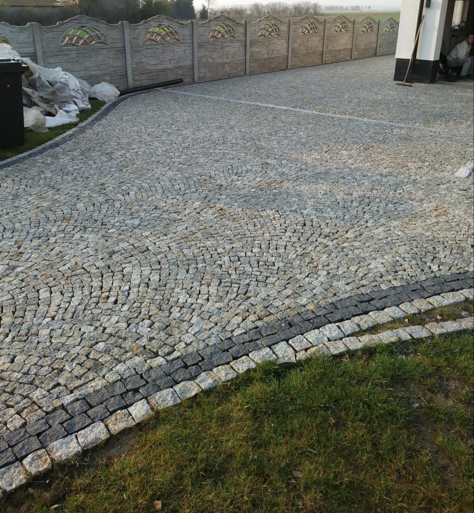 Podjazd wyłożony drobną, szarą kostką z ciemnoszarą obwódką, widoczne trawnik i betonowe ogrodzenie z dekoracyjnymi elementami.