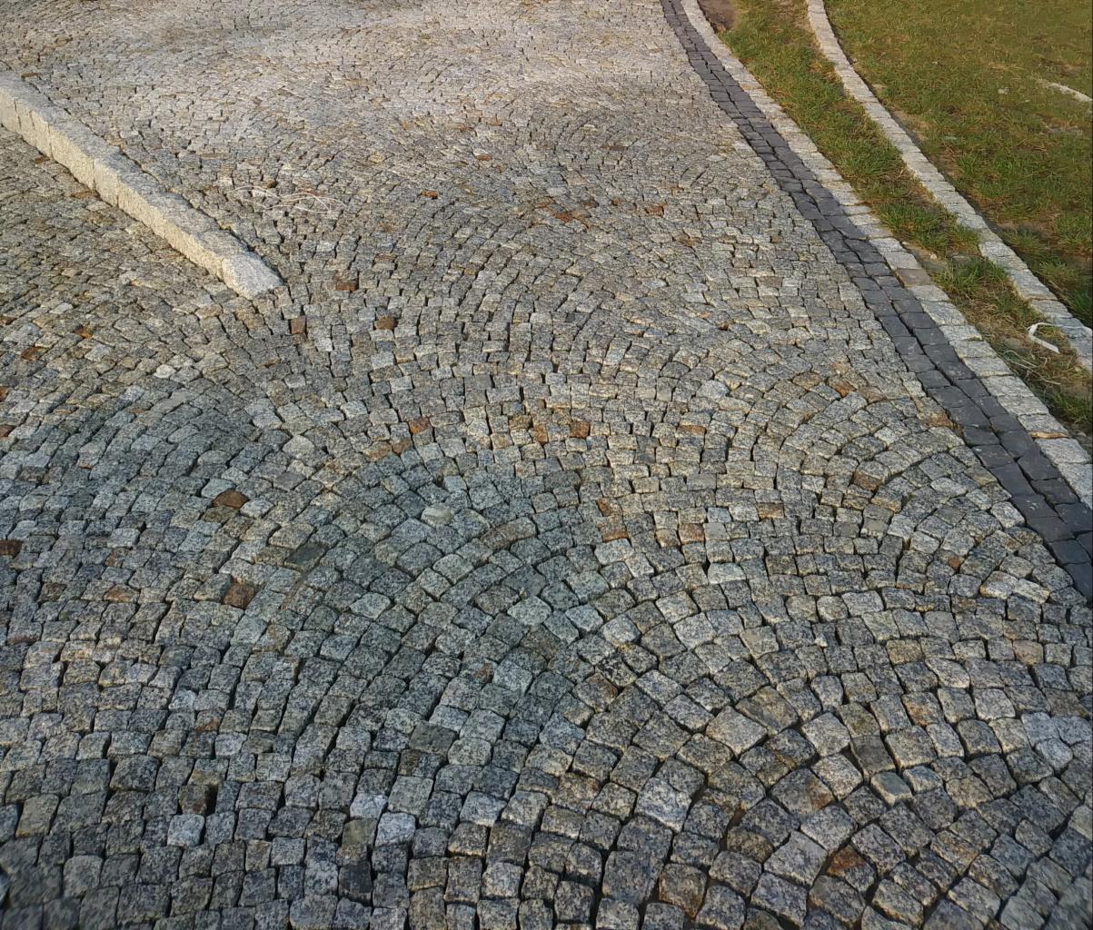 Szara, łukowato ułożona nawierzchnia z drobnej kostki, obrzeże z kostki w kolorze antracytowym oraz trawnik z boku.