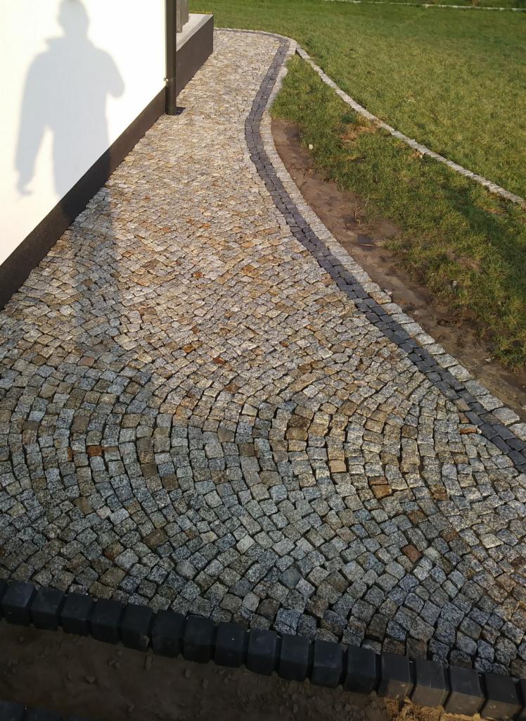 Ścieżka z drobnej kostki w odcieniach szarości i brązu, ułożona w łukowaty wzór, obrzeża z ciemnej kostki, trawnik w tle, cień osoby na ścianie budynku.