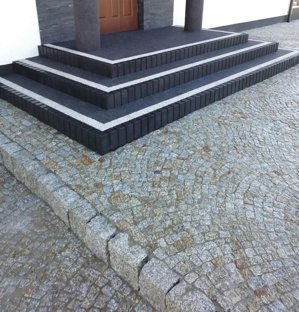 Schody wejściowe do domu wyłożone ciemną kostką z jasnymi fugami, połączone z chodnikiem z nieregularnej, szarej kostki. Widoczne dwa słupy podtrzymujące zadaszenie nad wejściem.