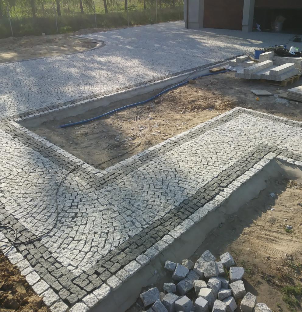 Fragment układanej nawierzchni z kostki, widoczne etapy prac: ułożony fragment z jasnej kostki w kontrastowym obramowaniu z ciemniejszej, przygotowane podłoże, zapas kostki w rogu, w tle fragment...