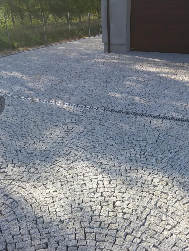 Ułożona nawierzchnia z drobnej, szarej kostki w kształcie wachlarza, z widocznym odpływem liniowym i fragmentem bramy garażowej w tle.