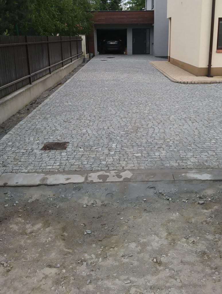 Podjazd do garażu wyłożony szarą kostką, widoczne ślady świeżych prac brukarskich w postaci ziemi i zaprawy. Zakamuflowana studzienka kanalizacyjna wkomponowana w nawierzchnię.
