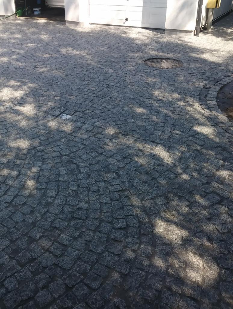 Nawierzchnia z ciemnoszarej kostki ułożonej w łukowaty wzór, z widocznym włazem kanalizacyjnym i cieniem drzewa, prowadząca do białego garażu.