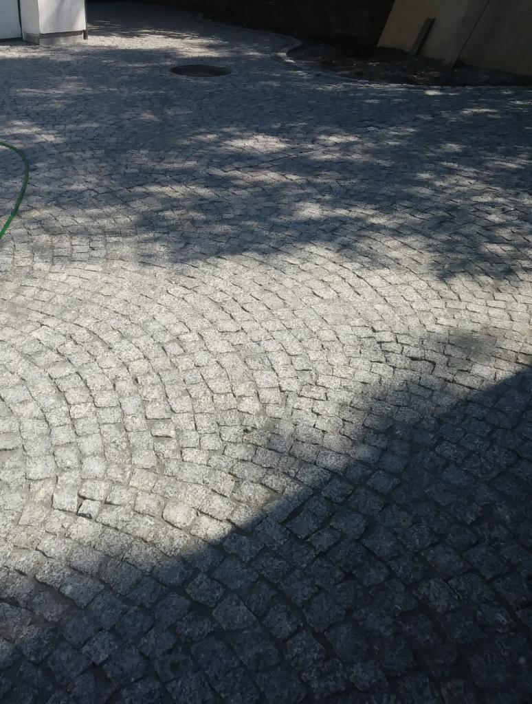 Szara nawierzchnia z kostki ułożonej w łukowaty wzór, częściowo zacieniona, z widocznym włazem kanalizacyjnym i fragmentem węża ogrodowego.