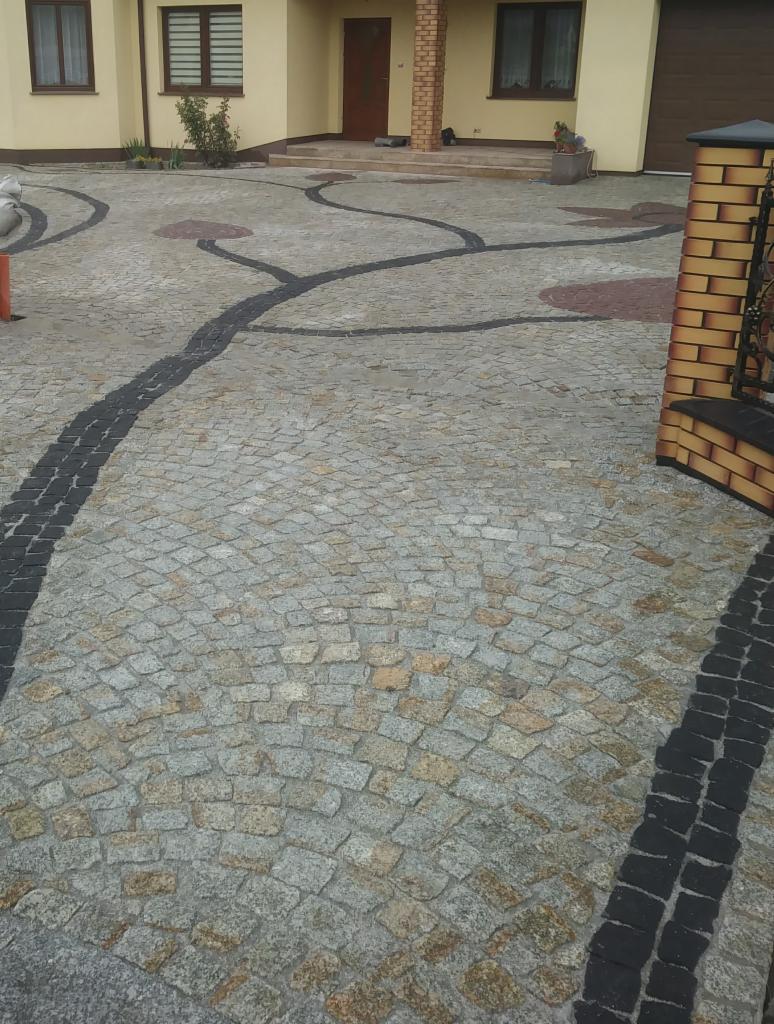 Ułożona nawierzchnia z drobnej, szaro-złotej kostki z dekoracyjnymi, ciemnymi pasami tworzącymi wzór przypominający gałęzie drzewa, prowadząca do domu z beżową elewacją i brązowymi oknami.