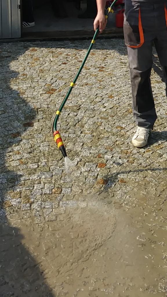 Czyszczenie powierzchni z kostki za pomocą węża ogrodowego z dyszą, widoczne zabrudzenia i woda na powierzchni, pracownik w ubraniu roboczym.